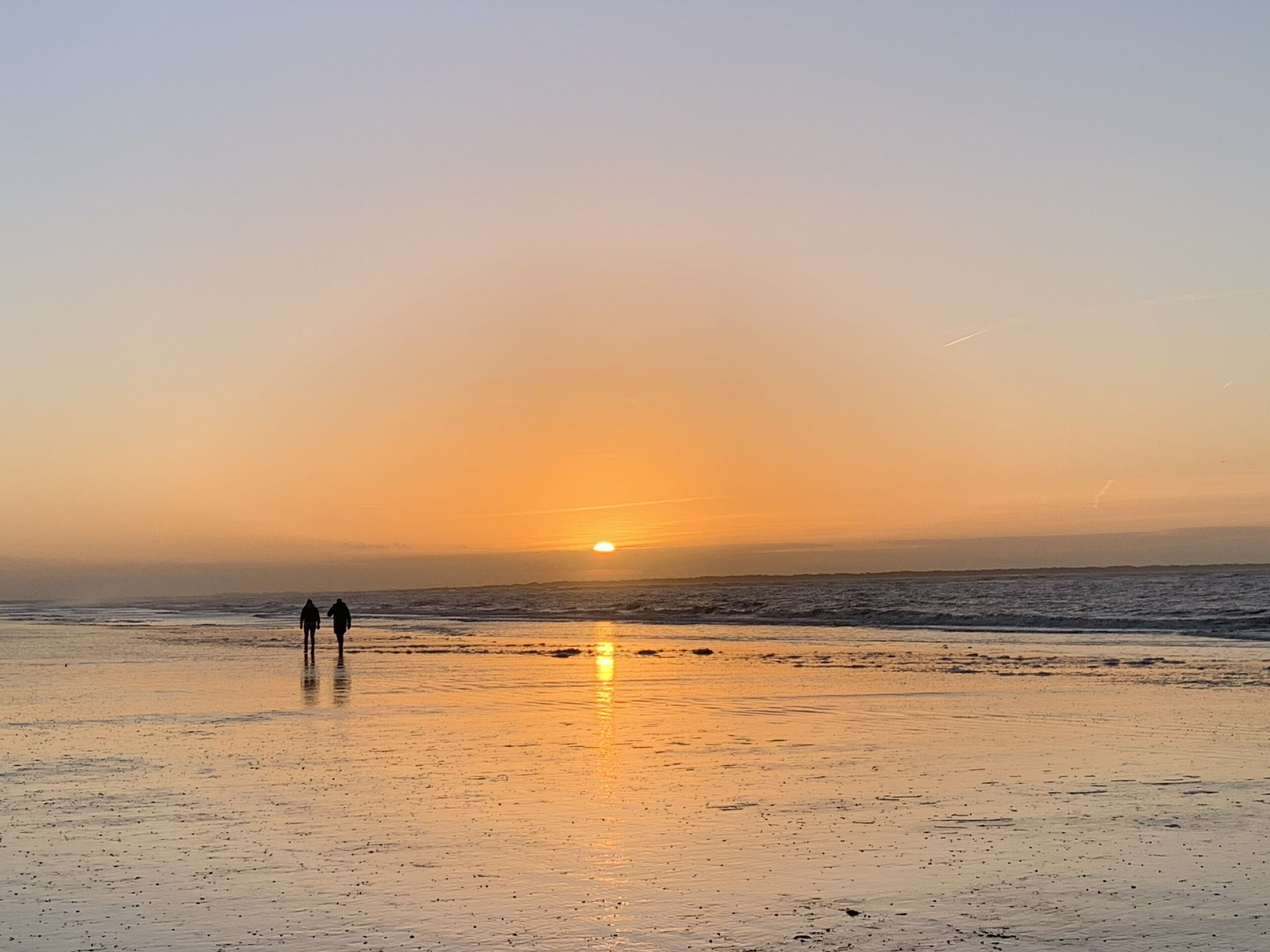 A beach at sunset with two people walking on the sand.