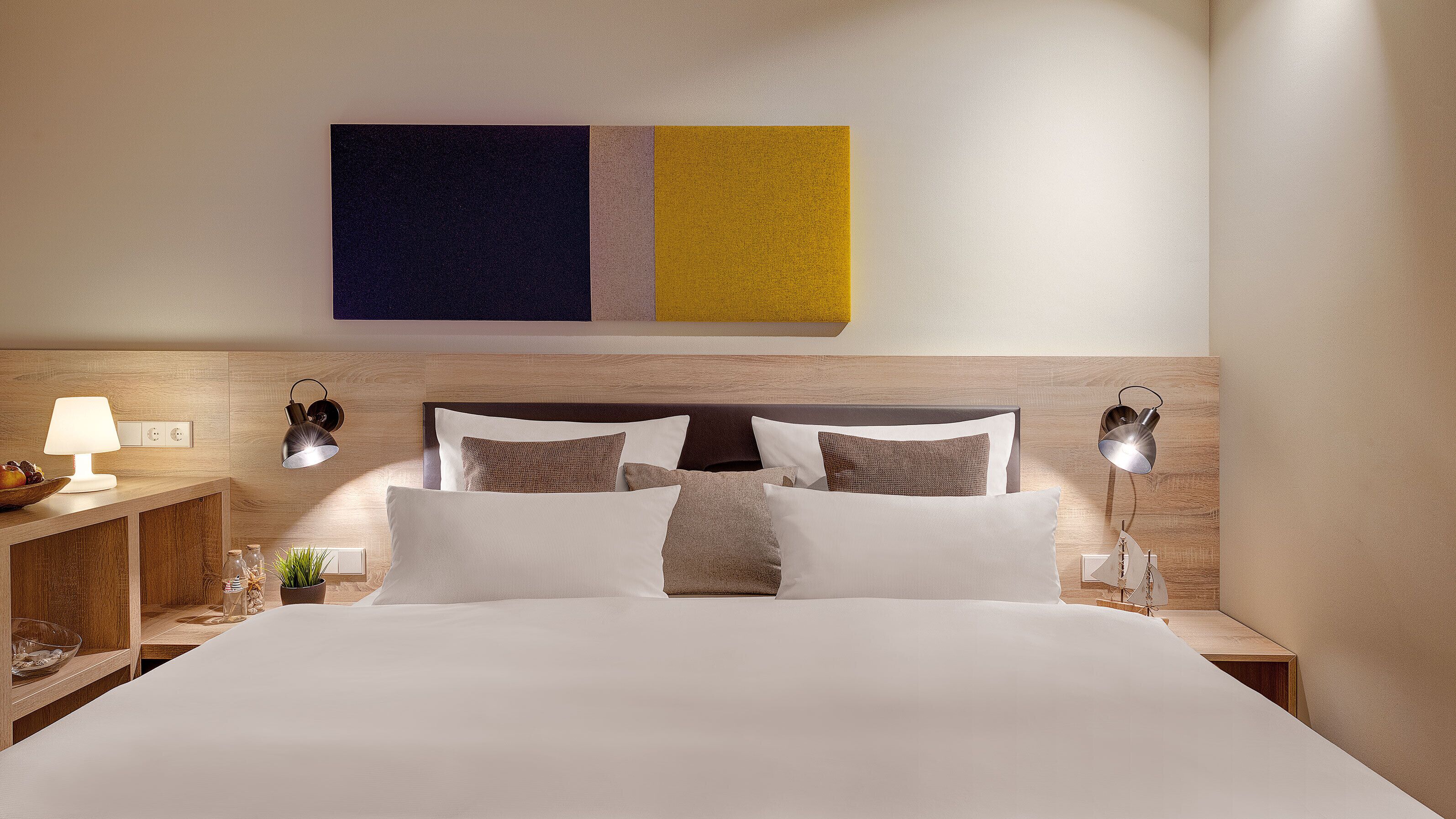 A neatly made bed with white linens and multiple pillows, a bedside table with a lamp, and a wall-mounted artwork above the bed.