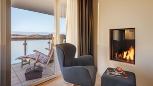 A balcony with a view of the water, a fireplace, a chair, and a small table with a book and a glass.