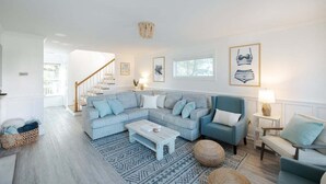 A spacious living room with a light blue sectional sofa, a coffee table, and a patterned rug. A staircase leads to another room, and there are framed pictures on the walls. - New! Close to Beach w Hot Tub! (Carolina Beach)