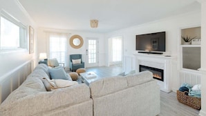 A modern living room with a sofa set, a television, a fireplace, and a large window. - New! Close to Beach w Hot Tub! (Carolina Beach)