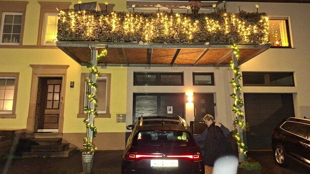 A building with a covered entrance, a car parked in front, and a person standing by the entrance. - Urgemütliche Ferienwohnung, Offenes Spitzdach Nähe Weinanbaugebieten (Salmtal)
