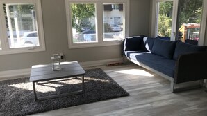 Un salon avec un canapé bleu foncé, une petite table basse en bois et un tapis gris. Trois fenêtres offrent une vue sur l’extérieur.