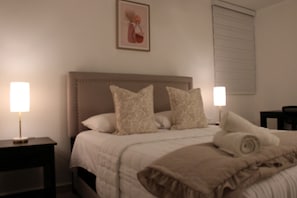 Una cama bien hecha con cabecera, dos lámparas de noche y un cuadro en la pared.