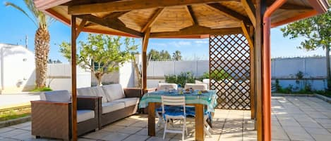 Eine Terrasss mit Holz-Pergola, Tisch mit Stühlen, einer Couch und einem Palmenbaum.