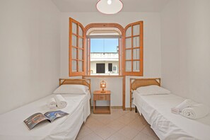 A hotel room with two single beds, a small bedside table with a lamp, a window with wooden shutters, and a view of the outside.