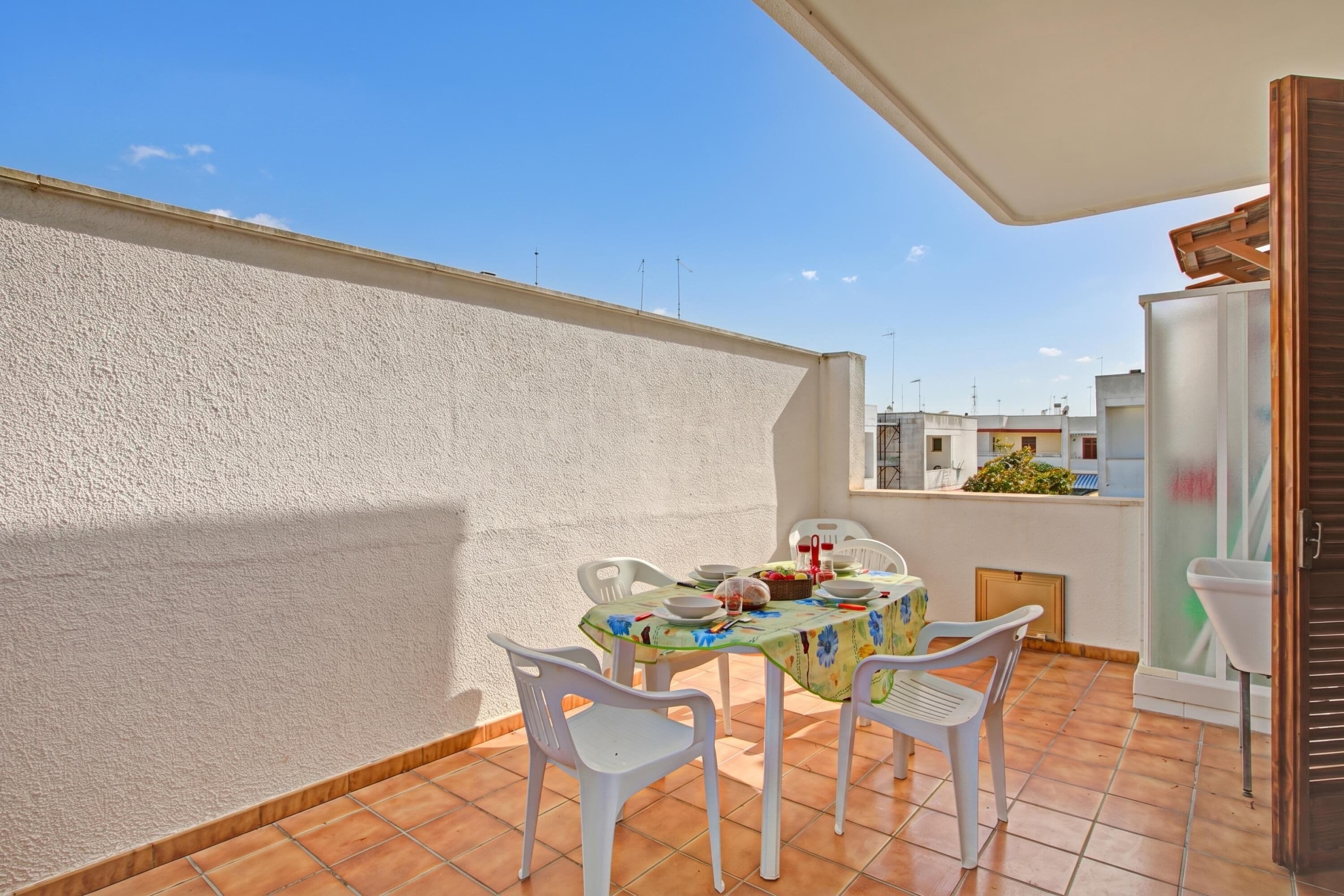 Un balcon avec une table dressée pour le repas, des chaises en plastique blanches et un mur blanc.