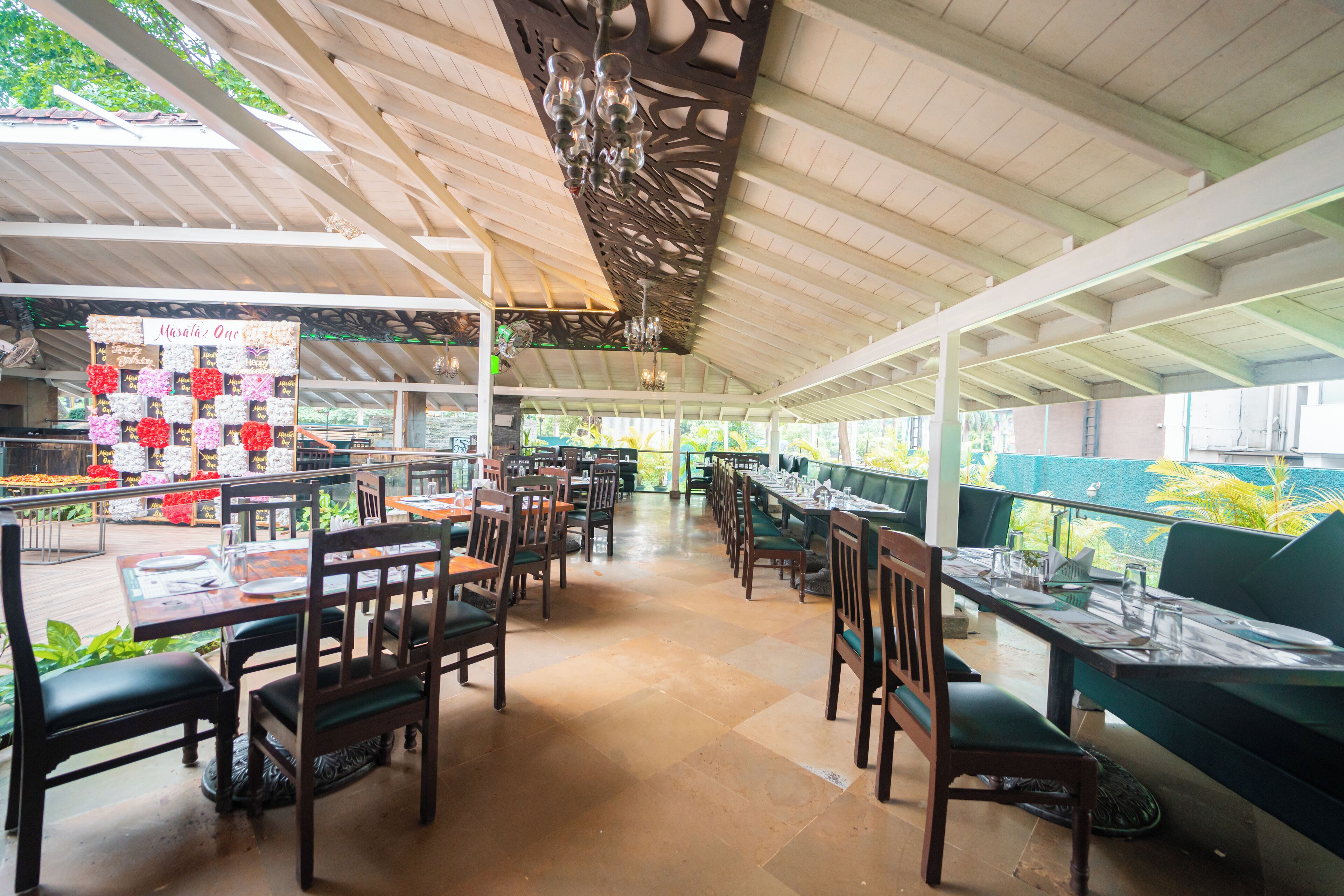 Un amplio espacio exterior para comer con mesas y sillas de madera, techo decorativo y vista a la vegetación.