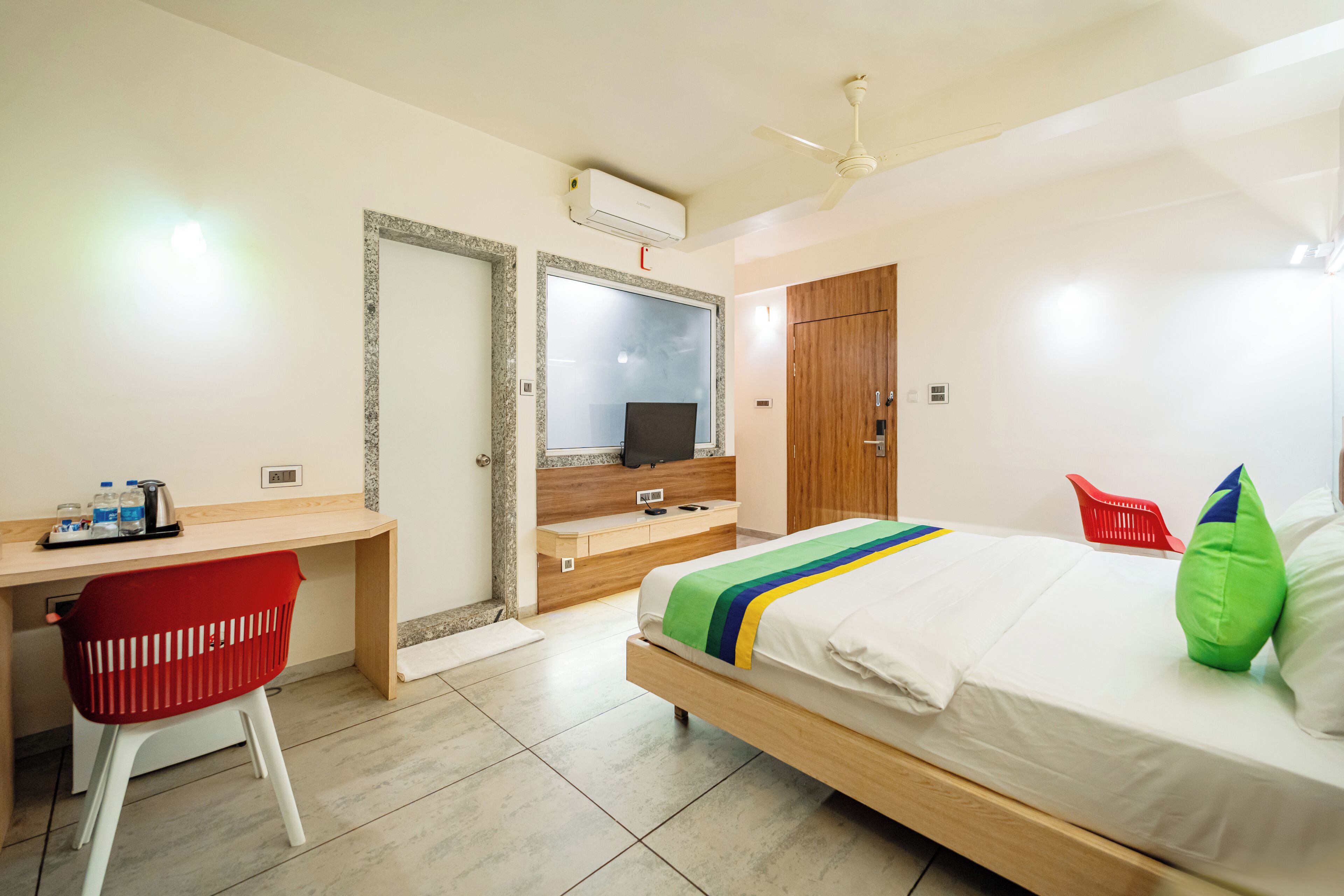 A hotel room with a bed, a desk with a chair, a wardrobe, and a mirror.