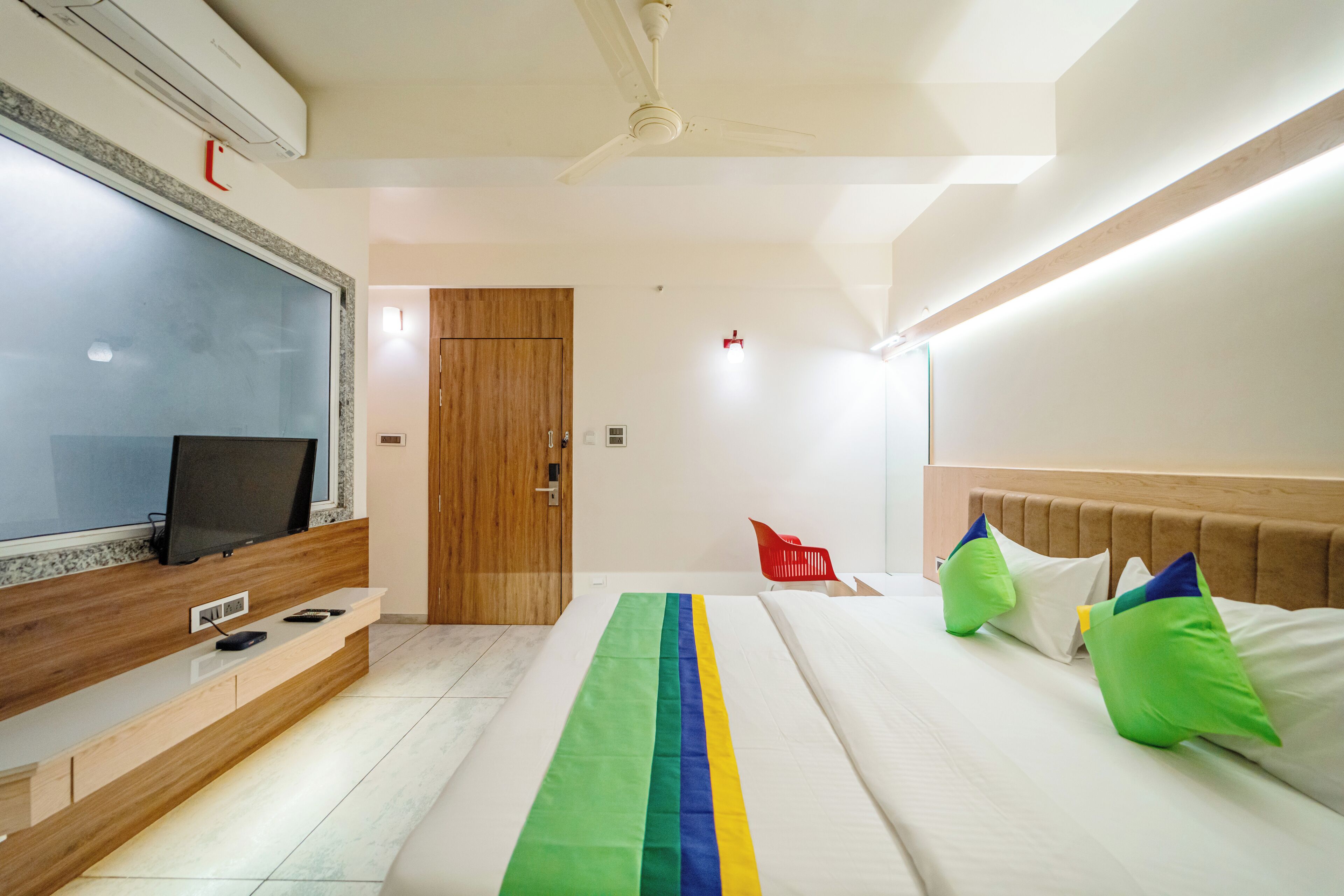 Una habitación de hotel moderna con un televisor de pantalla plana grande, una mesa consola de madera, una cama con almohadas de colores y un ventilador de techo.