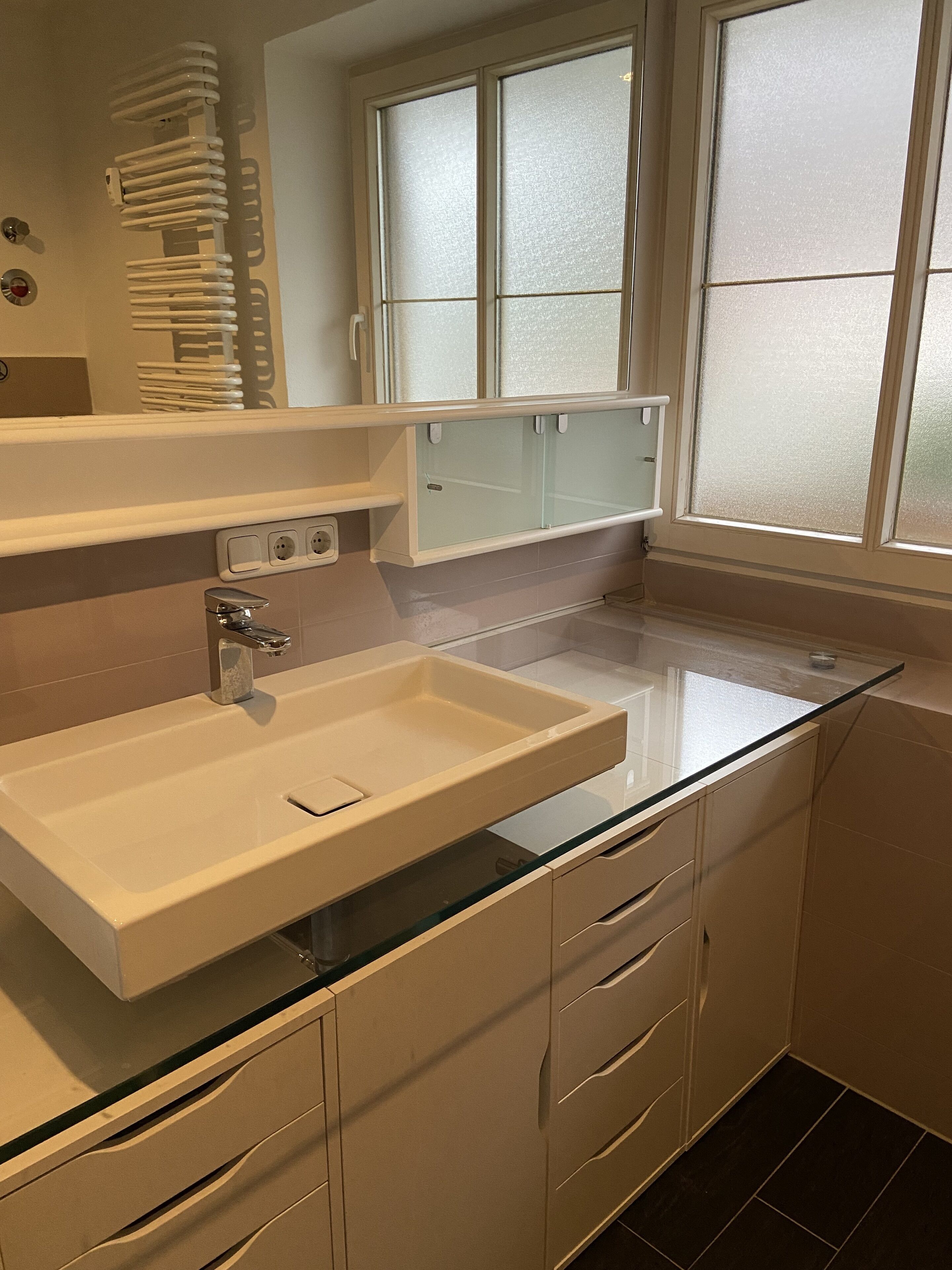 A modern bathroom with a glass-enclosed sink, white cabinetry, and frosted glass windows.