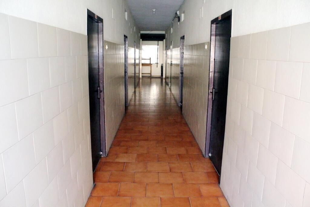 Un couloir aux murs recouverts de carreaux blancs et au sol carrelé de carreaux bruns, qui mène à une porte au fond.