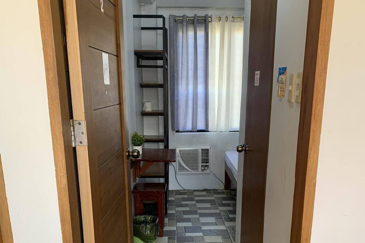 Un pasillo estrecho con suelo de baldosas, un felpudo verde, una pequeña mesa con una planta, un estante y una cama con una ventana y cortinas al fondo.