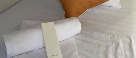 Una cama bien hecha con sábanas blancas, una almohada marrón y una toalla blanca enrollada con la etiqueta 'GUEST KIT'.