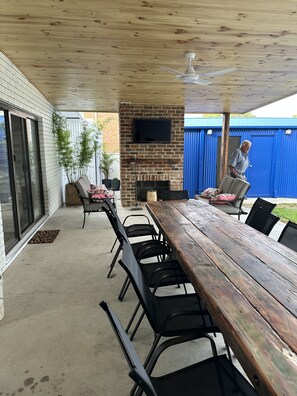 Eine Terrasse im Freien mit einem Holztisch und Stühlen, einem an einer Ziegelwand montierten Fernseher und einem Deckenventilator.
