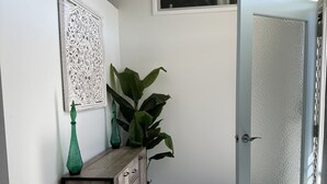 Un couloir avec une console en bois, un vase vert, une grande plante et une porte en verre dépoli.