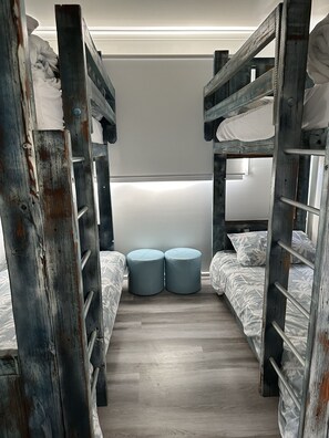 Ein Mehrbettzimmer mit zwei runden Hocker in der Mitte, Holzbetten links und rechts sowie einer schlichten weißen Wand im Hintergrund.