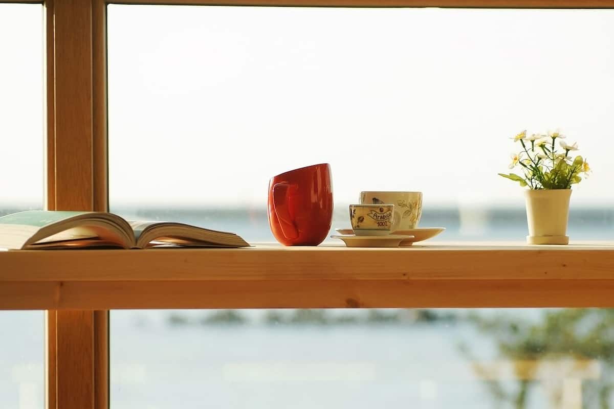 Ein Holzregal mit einem Buch, einer roten Tasse, einer Kaffeetasse und einem kleinen Topfpflanzen.
