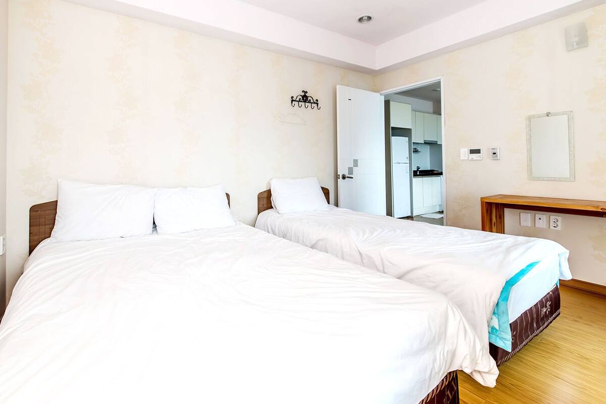 Ein Hotelzimmer mit zwei Einzelbetten, einem Holzkopfstück, einem kleinen Nachttisch, einem Spiegel und einer Tür zu einer Küchenzeile.