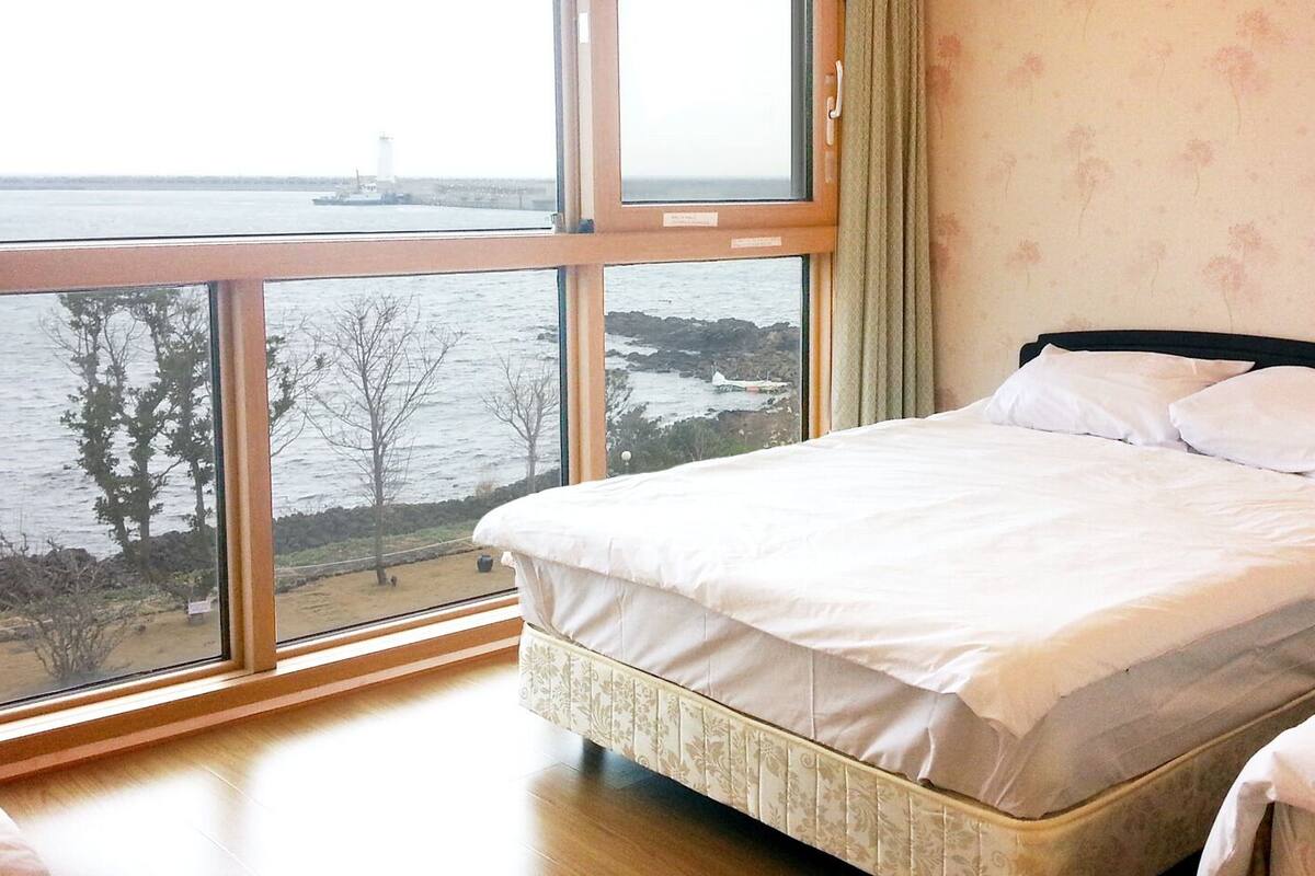 Ein Schlafzimmer mit großem Fenster mit Blick auf ein Gewässer und einen entfernten Leuchtturm.