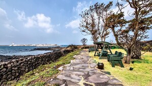 A stone pathway leading to a grassy area with a swing set and picnic tables, overlooking a rocky coastline with a lighthouse in the distance. - Aerang and Baebijang's House (Jeju City)