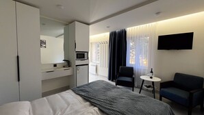 Une chambre d’hôtel avec un lit, un bureau avec un ordinateur, une chaise, une petite table avec une bouteille et des verres, un téléviseur fixé au mur et une fenêtre avec des rideaux.