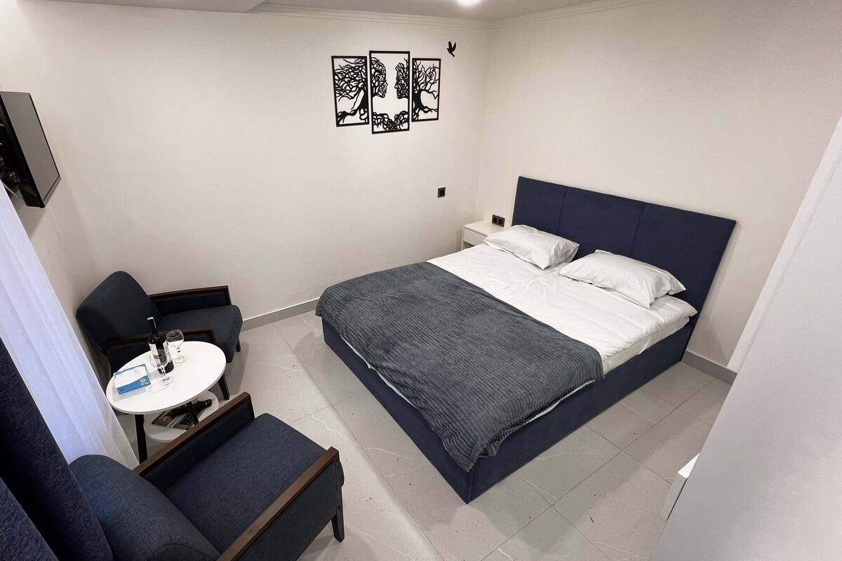 A compact hotel room with a single bed, a small round table with a bottle and glasses, a chair, and a TV mounted on the wall.