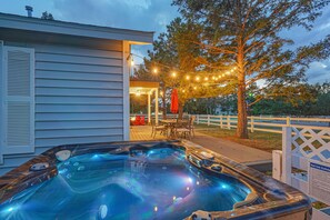 Un jacuzzi con terraza de madera y luces colgantes, rodeado por una valla blanca y árboles.
