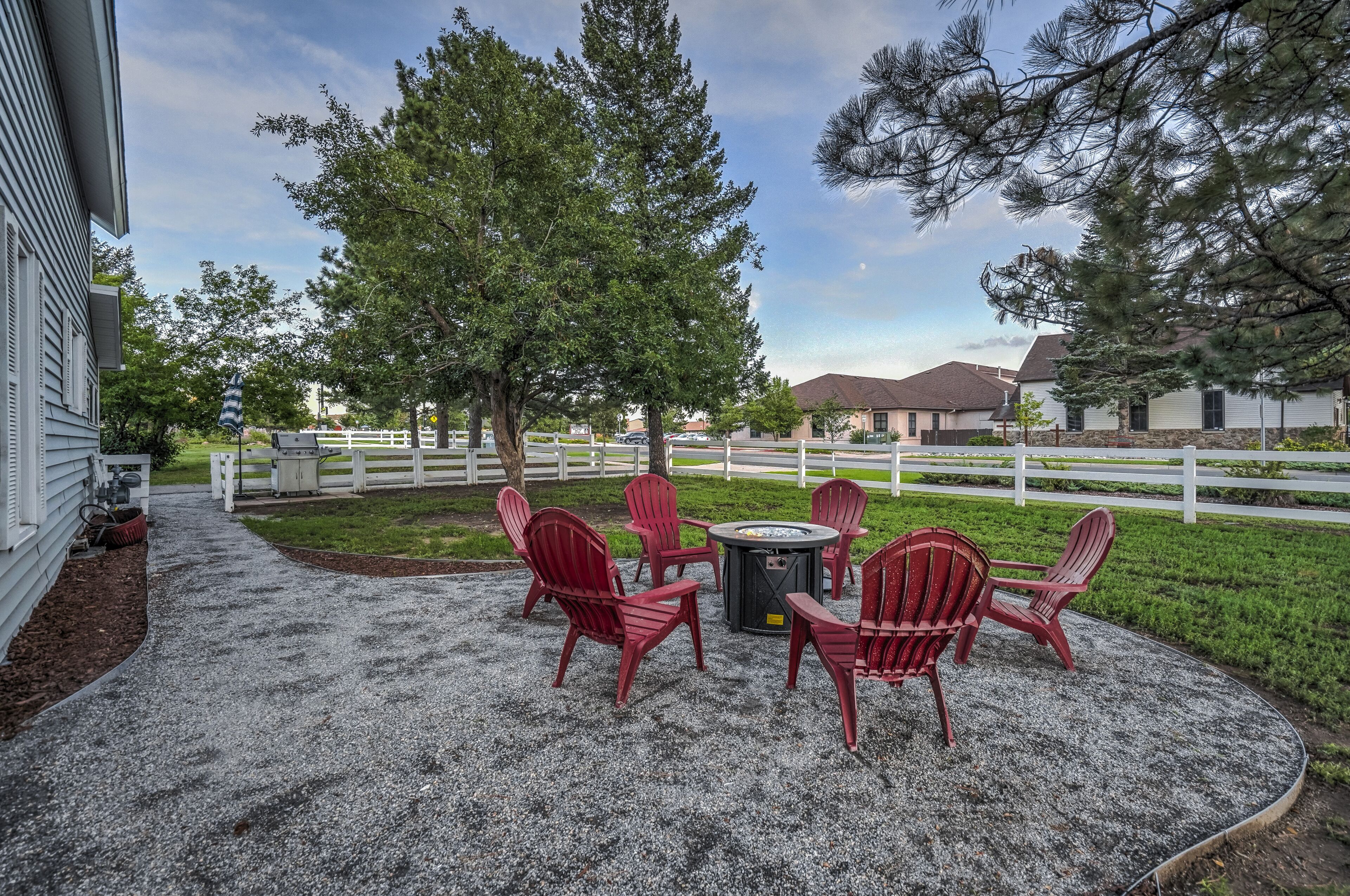 A patio with red Adirondack chairs and a fire pit, surrounded by a white fence and trees.