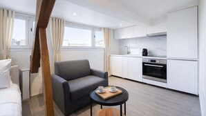 Une chambre d’hôtel moderne avec un canapé gris, une petite table ronde sur laquelle se trouve un plateau, une chaise en bois, un lit, une kitchenette équipée d’une cuisinière et d’un four, et de grandes fenêtres avec des rideaux.