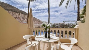 A balcony with white plastic chairs and a table set for two, a vase with flowers, and a view of a coastal town. - Castle Harbour 3 (Los Cristianos)