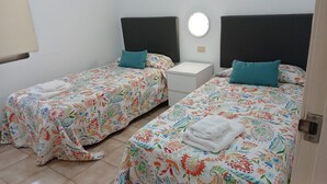 A twin room with two beds, each with a floral bedspread and teal pillows, a bedside table, and a round wall light. - Castle Harbour 3 (Los Cristianos)