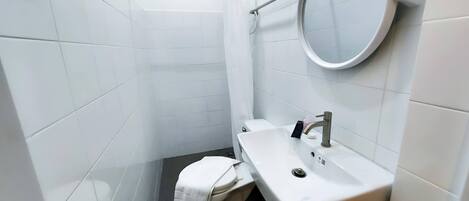 A small, modern bathroom with white tiles, a round mirror, a toilet, and a sink.