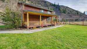 A log cabin with a porch, surrounded by a grassy yard and a hillside.