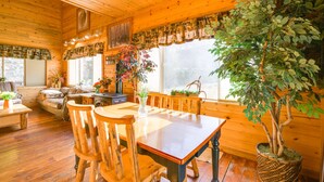 A wooden cabin interior with a dining table, chairs, a sofa, and a large window with curtains.