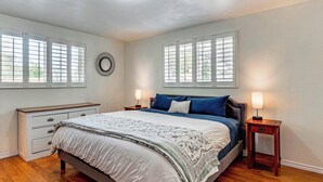 Une chambre à coucher comprenant un lit, deux tables de chevet, une commode et une fenêtre avec des stores.