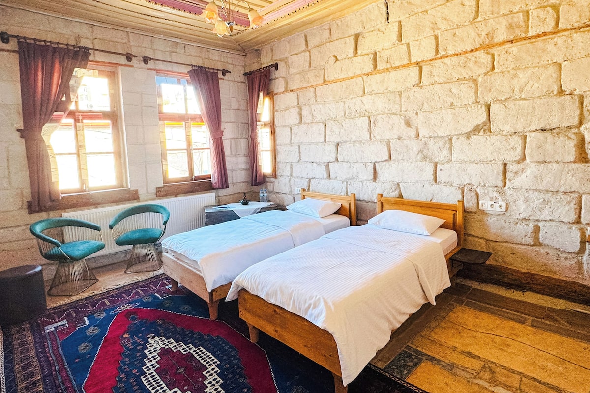 A room with two wooden beds, a radiator, a window with curtains, a chair, and a patterned rug.