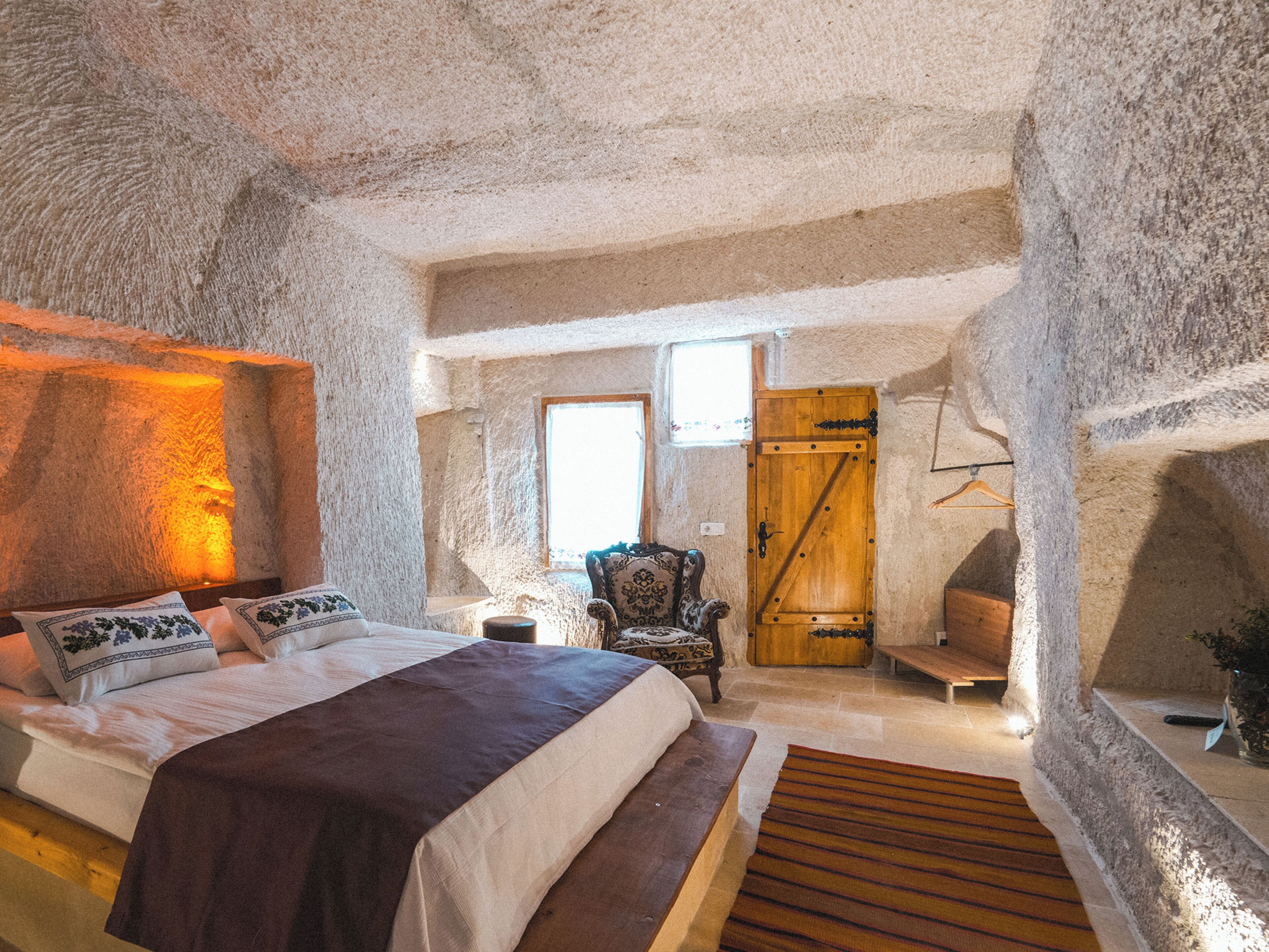 A bedroom with a stone wall, a bed, a wooden bench, a chair, and a wooden door.