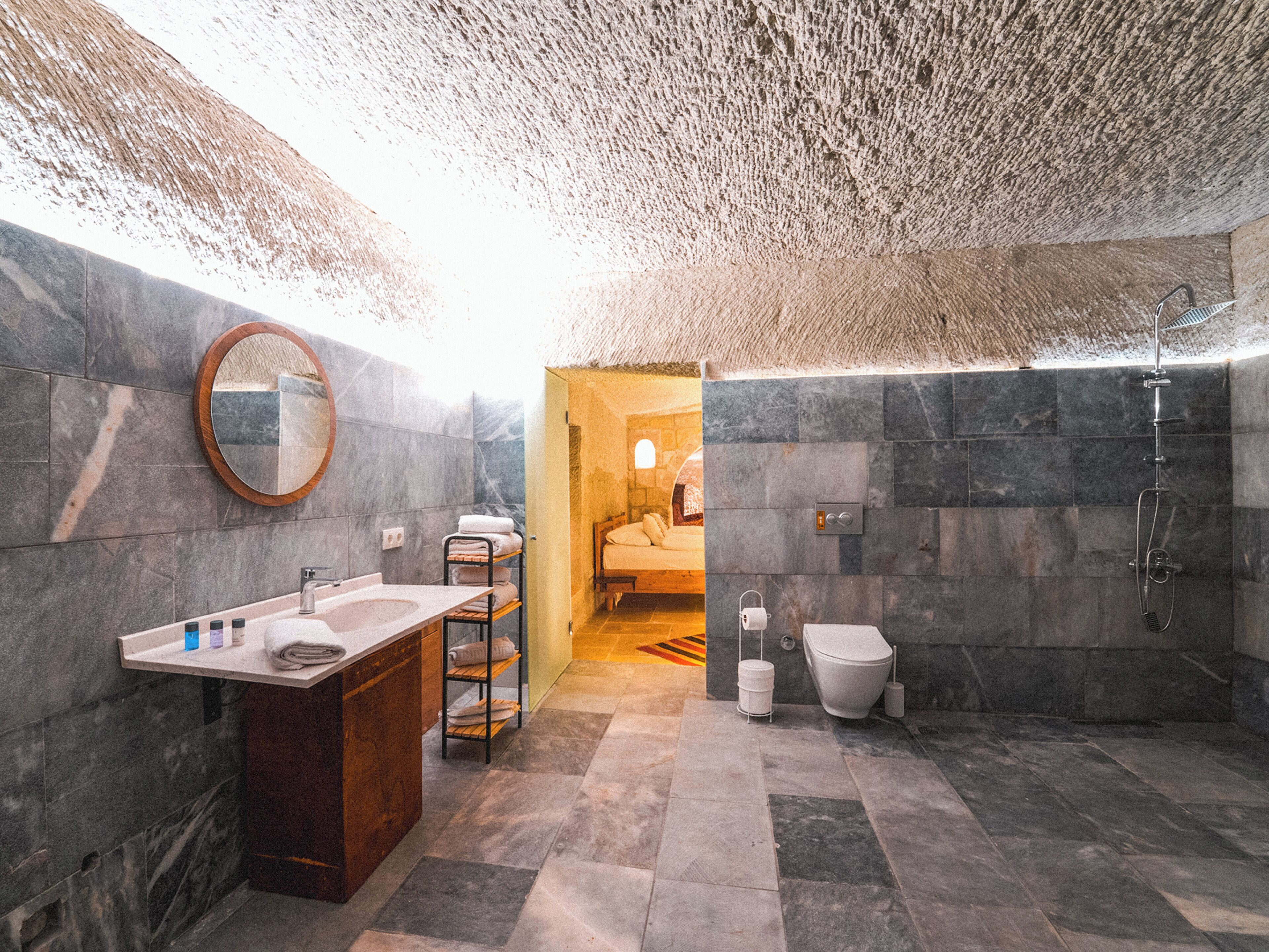 A modern bathroom with a marble sink, a wooden vanity, a round mirror, a towel rack, a shower, and a toilet.