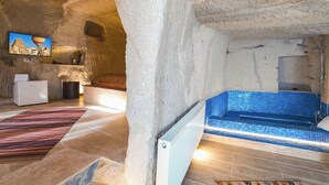 A stone-walled interior with a blue-tiled hot tub, a white refrigerator, and a television displaying a mountainous landscape. - MY CAVE SUITE (Nevsehir)