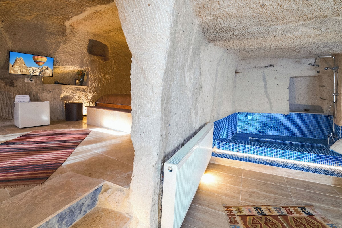 A stone-walled interior with a blue-tiled hot tub, a white refrigerator, and a television displaying a mountainous landscape.