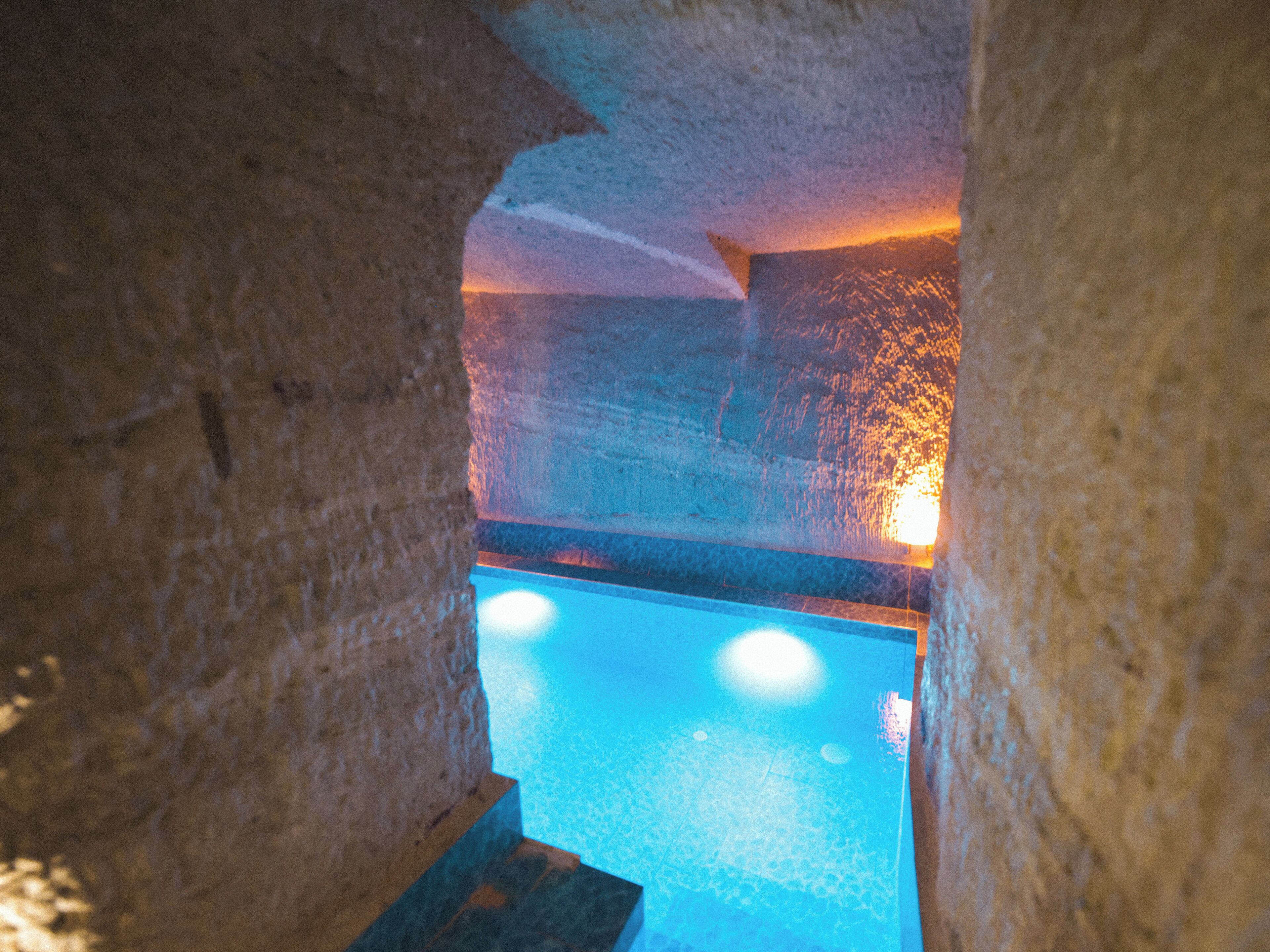 Un área de piscina con paredes de piedra, piscina azul cristalina y una fuente de luz cálida al fondo.