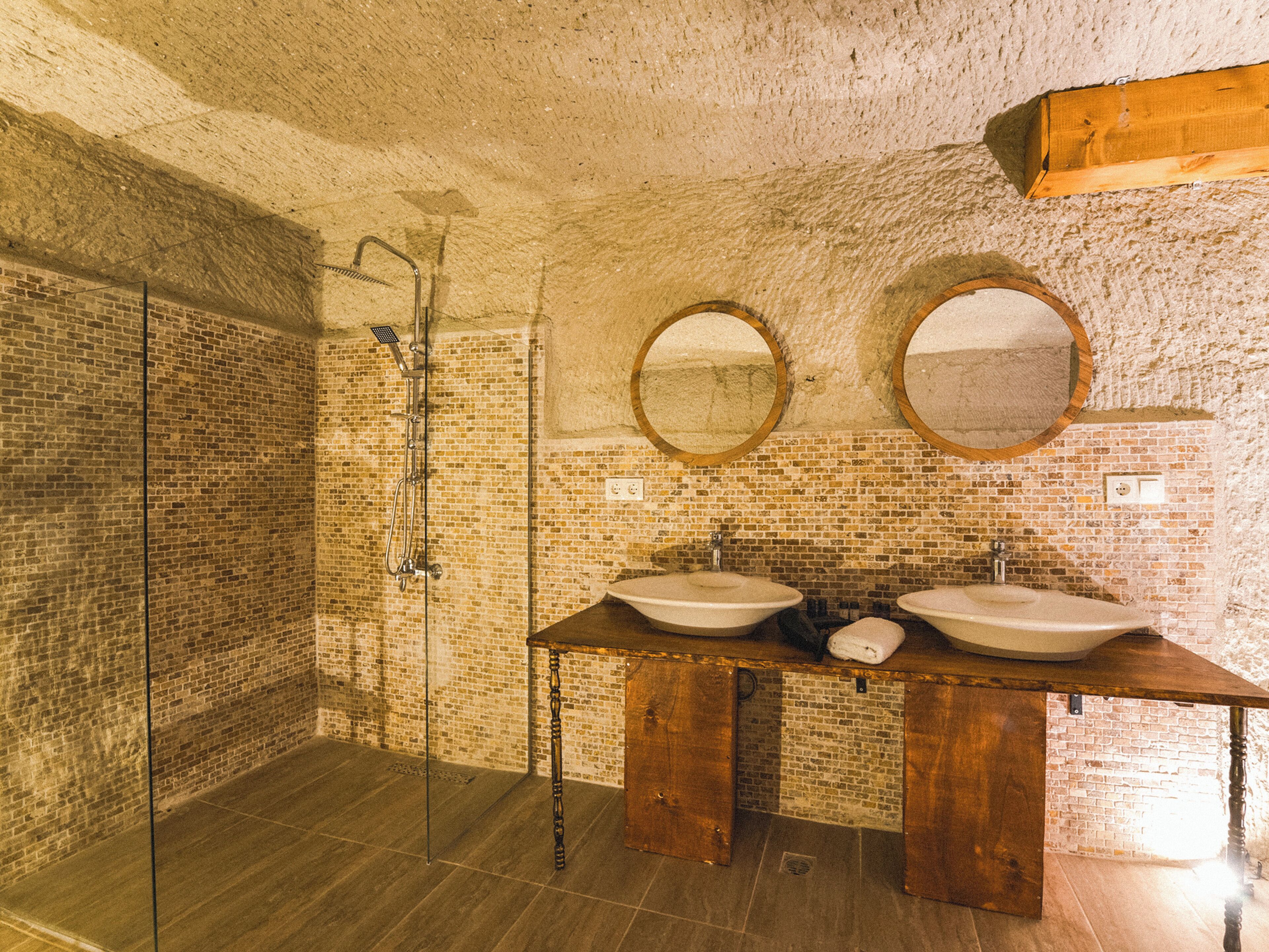 Casa de banho com parede de tijolo, cabine de duche em vidro e duas pias com espelhos redondos.