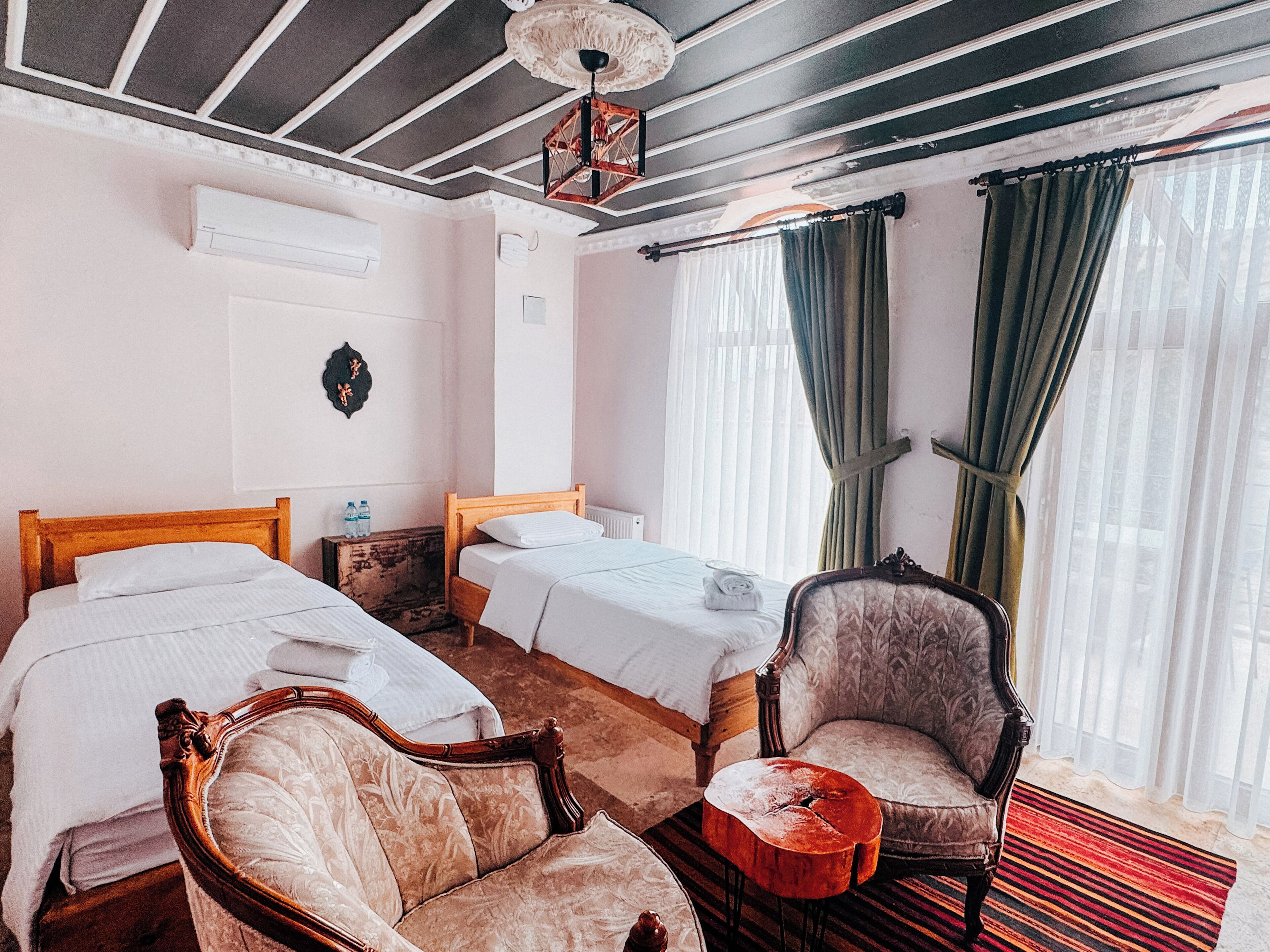Habitación de hotel con dos camas, cabecero de madera, una silla, una mesita y una ventana con cortinas.