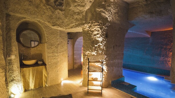 A spa room with a stone wall, a round mirror, a sink, a wooden bench, and a small pool. - MY CAVE SUITE (Nevsehir)