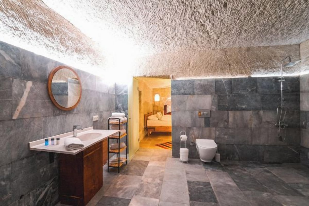 A modern bathroom with a marble sink, a wooden vanity, a shower, and a visible bedroom in the background.