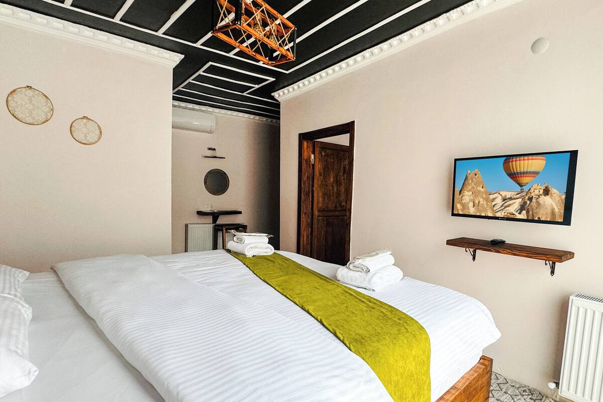 A hotel room with a large bed, a wooden headboard, a green throw blanket, a wall-mounted TV, and a painting of a hot air balloon.