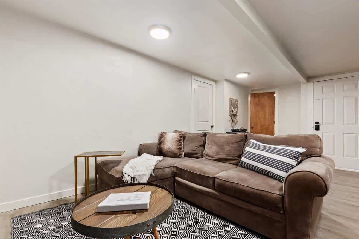 A living room with a brown sectional sofa, a round coffee table, and a patterned rug. There are two white doors and a wooden door in the background.