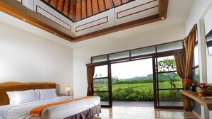 A spacious bedroom with a large bed, a view of a lush landscape through floor-to-ceiling windows, and a wooden ceiling design. - Lumbung Asri Cottage (Mengwi)