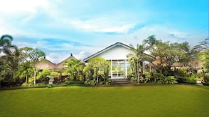 A white building with a glass entrance, surrounded by lush greenery and a well-maintained lawn. - Lumbung Asri Cottage (Mengwi)
